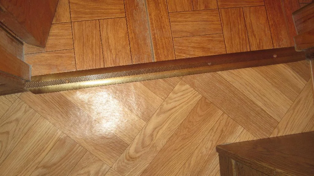 Металлический порог установлен в дверной проем туалета. Плитка и линолеум коричнево-бежевого цвета. Дверной порог темно-коричневый.