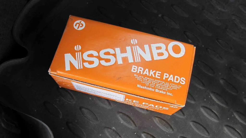 Тормозные колодки Nisshinbo PF-3548 в заводской коробке лежат на автомобильном коврике.